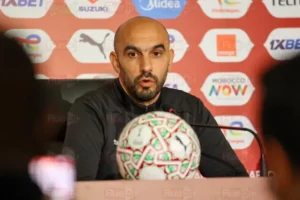 Walid Regragui speaks at a press conference, with a football adorned in Moroccan colours prominently in the foreground and a branded backdrop.