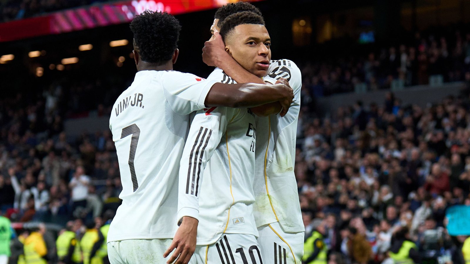 Kylian Mbappé, met rugnummer 10, viert met Vinícius Jr. na een doelpunt, omringd door opgewonden fans in het stadion.