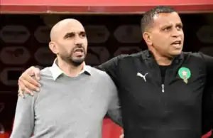Walid Regragui et son entraîneur adjoint s'enlacent, tous deux vêtus de l'habit de l'équipe, devant un fond de football marocain, dégageant de la camaraderie.