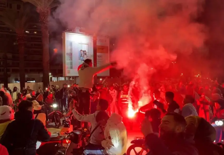Moroccan fans celebrate in a packed street, lit by red flares and surrounded by smoke, showcasing their passion for football.
