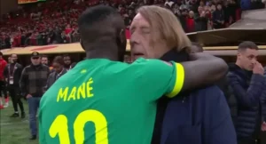 Sadio Mané embrasse l'entraîneur du Sénégal après un match, illustrant un moment de camaraderie, avec des supporters en arrière-plan.