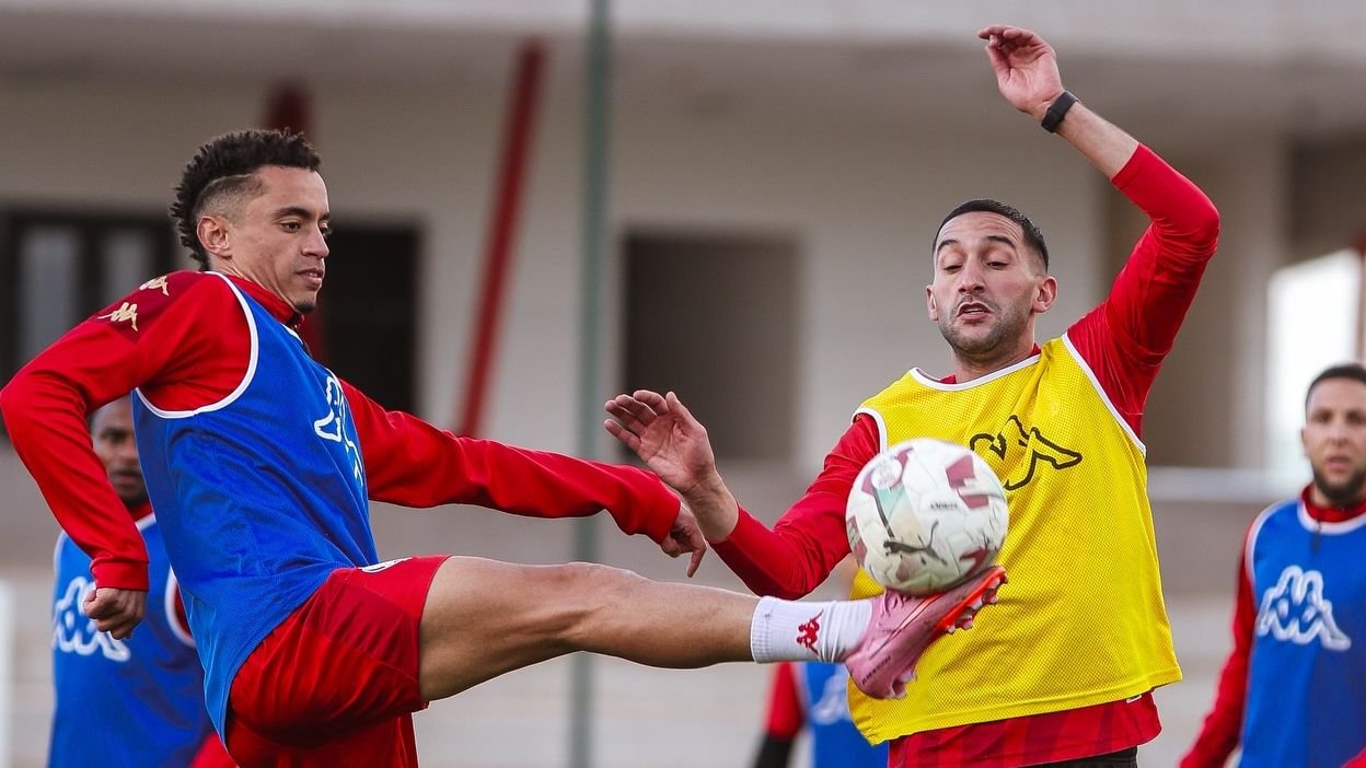 Wydad Casablanca-spelers tijdens training, gekleed in rode en blauwe tenues, terwijl een speler een trap probeert te geven en een ander zich klaarmaakt om de bal te onderscheppen.