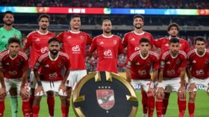تشكيلة فريق الأهلي بقمصان حمراء تلتقط صورة مع كأس قبل المباراة، على خلفية ملعب نابض بالحياة.
