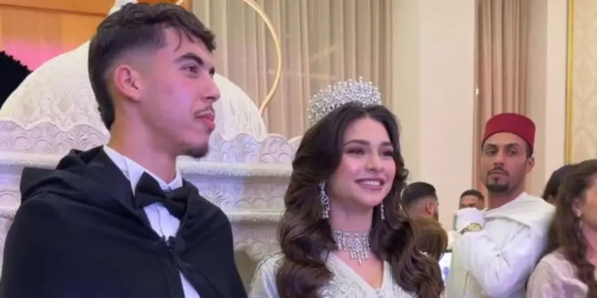 Zabiri in formal attire stands beside a smiling woman in traditional dress and a crown, with guests and ornate decor in the background.
