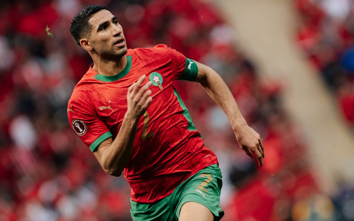 Achraf Hakimi sprinte dans le maillot national rouge et vert vif du Maroc pendant un match, avec des fans enthousiastes en arrière-plan.