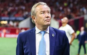 أدريان هيث، يرتدي بدلة زرقاء وربطة عنق فاتحة، يحدق بتأمل خلال المباراة بينما يظهر المشجعون مشوشين في الخلفية.