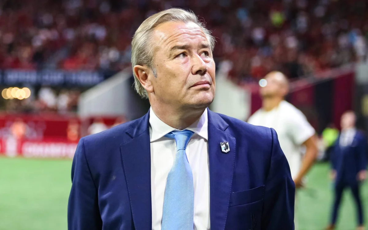 أدريان هيث، يرتدي بدلة زرقاء وربطة عنق فاتحة، يحدق بتأمل خلال المباراة بينما يظهر المشجعون مشوشين في الخلفية.