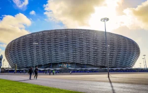 Modern stadium exterior with unique lattice design, surrounded by fans and bright blue sky, showcasing anticipation for the 2026 Africa Cup.