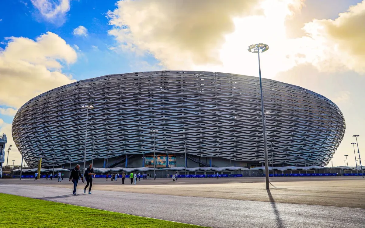 Modern stadium exterior with unique lattice design, surrounded by fans and bright blue sky, showcasing anticipation for the 2026 Africa Cup.