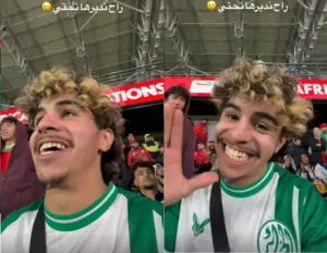 Un fan enthousiaste, vêtu d'un maillot vert et blanc, sourit et fait signe à la caméra, entouré d'autres supporters dans l'ambiance du stade.