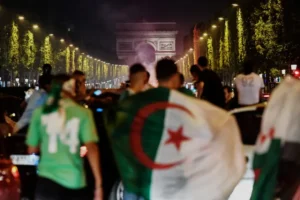 Algerijnse fans vieren in Parijs, zwaaiend met vlaggen nabij de Arc de Triomphe, verlicht door feestelijke lichten en rook in de nacht.