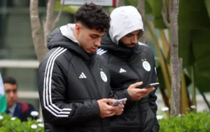 Algerijnse voetballers in zwarte teamjassen checken hun telefoons, omgeven door groen en fans op de achtergrond.