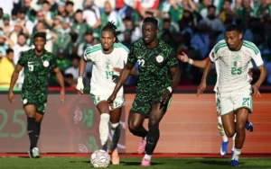 Les joueurs algériens en blanc affrontent un adversaire vêtu de vert lors de la Coupe d'Afrique 2025, illustrant une action de football intense.