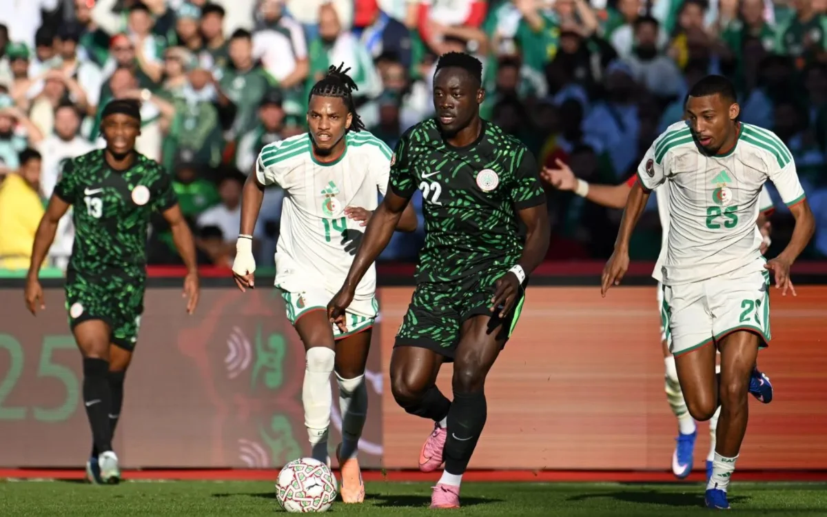 Les joueurs algériens en blanc affrontent un adversaire vêtu de vert lors de la Coupe d'Afrique 2025, illustrant une action de football intense.