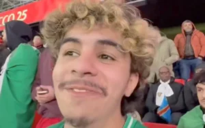 Glimlachende supporter in een groen shirt in het stadion van Rabat, met fans op de achtergrond, die een levendige wedstrijdsfeer toont.