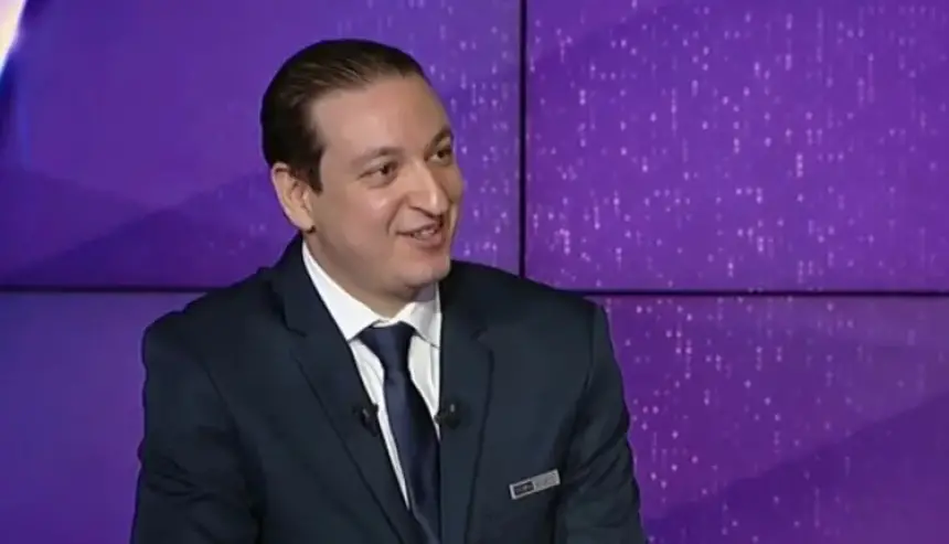 Un commentateur sportif souriant en costume marine avec une chemise blanche et une cravate noire est assis contre un fond violet, discutant de football.