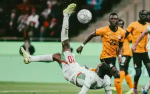 Ayoub El Kaabi exécutant une reprise acrobatique lors d'un match contre la Zambie, avec des joueurs en tenues verte et jaune sur fond de stade flou.