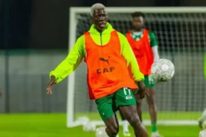 Player in a bright orange training bib with "CAF" logo, preparing to kick a football during a training session, with teammates in background.