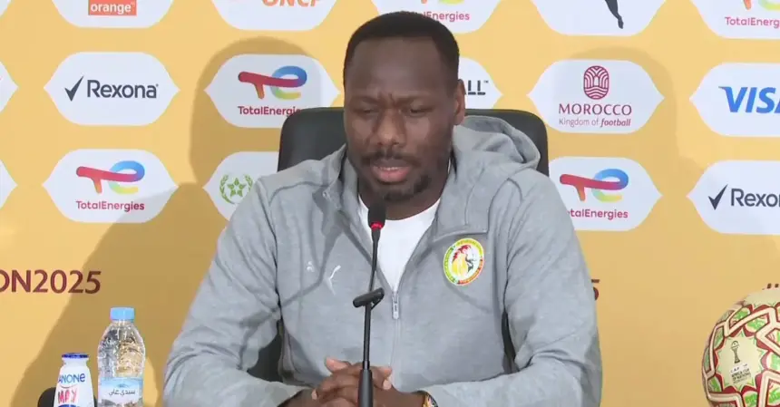 Senegal's football coach speaks at a press conference, wearing a grey jacket, set against a vibrant yellow backdrop with sponsorship logos.