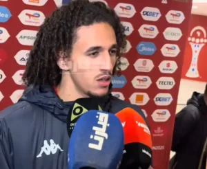 Moroccan footballer Amine Harit speaks to reporters in front of a branded backdrop, surrounded by microphones during a press interview.