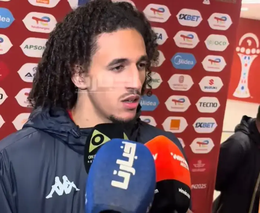 Moroccan footballer Amine Harit speaks to reporters in front of a branded backdrop, surrounded by microphones during a press interview.