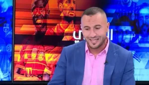 Analyste sportif portant un costume bleu et une chemise rose, avec des graphiques vibrants de joueurs de football en arrière-plan, discutant du football marocain.