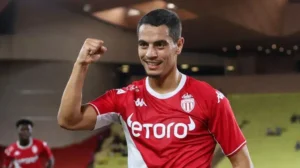 Wissam Ben Yedder célèbre dans un maillot rouge de Monaco, poing serré levé en triomphe, montrant un arrière-plan de stade vibrant.