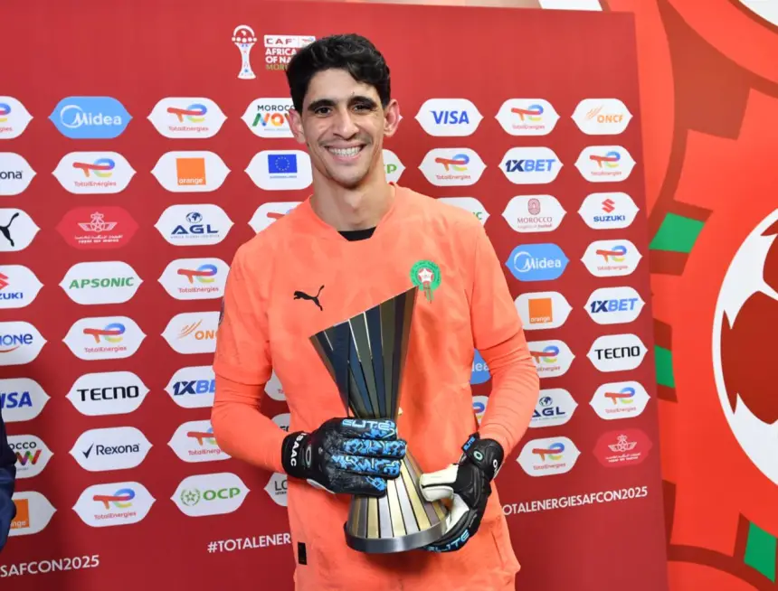 Yassine Bounou proudly holds the trophy, wearing a vibrant orange goalkeeper kit, set against a backdrop of the CAF AFCON 2025 branding.