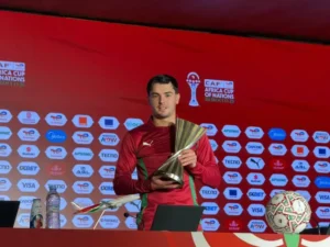 Brahim holding the CAF Africa Cup of Nations trophy, dressed in a maroon kit with green accents, against a red backdrop with sponsor logos.