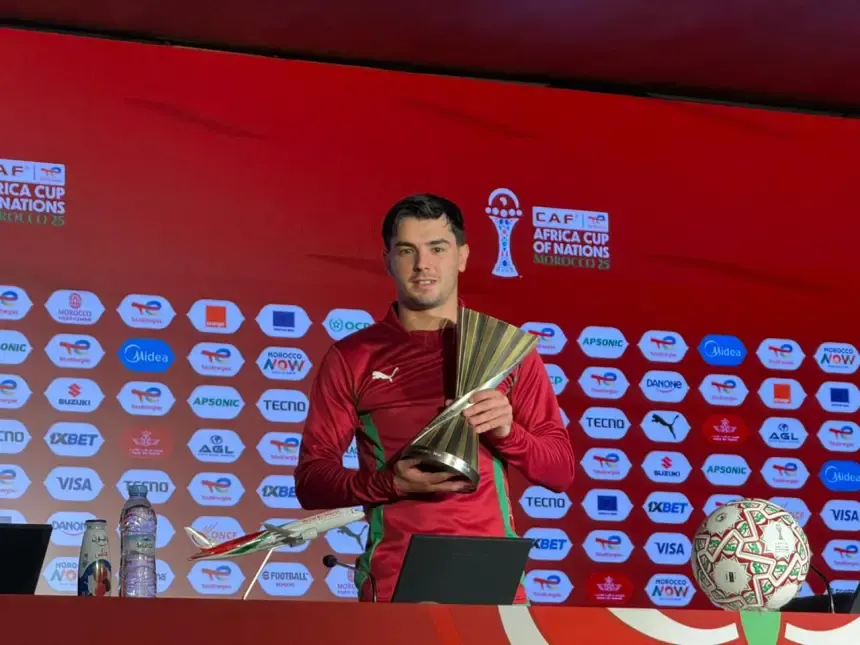 Brahim holding the CAF Africa Cup of Nations trophy, dressed in a maroon kit with green accents, against a red backdrop with sponsor logos.