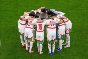 Les joueurs marocains, vêtus de maillots blancs avec des accents rouges et verts, se rassemblent en cercle, renforçant l'esprit d'équipe avant un match.