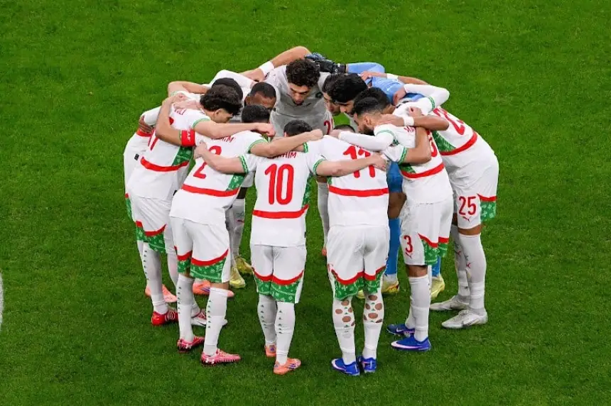 Les joueurs marocains, vêtus de maillots blancs avec des accents rouges et verts, se rassemblent en cercle, renforçant l'esprit d'équipe avant un match.
