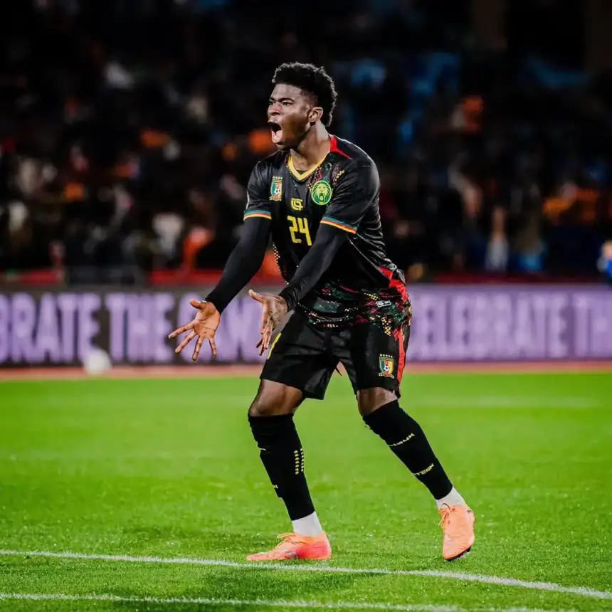 Joueur camerounais célébrant avec passion sur le terrain, vêtu d'un maillot noir aux motifs vibrants, dans une atmosphère de stade animée.