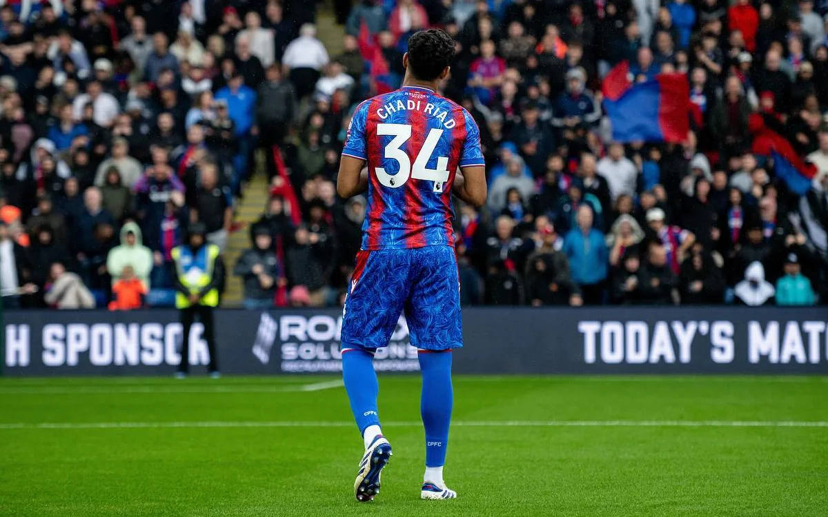 Chadi Riadi in Crystal Palace's vibrant blue and red kit, walking towards a cheering crowd during a match at Selhurst Park.