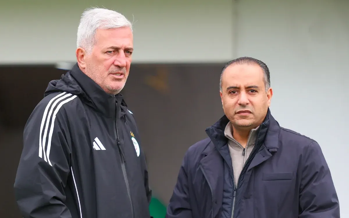 L'entraîneur algérien et son adjoint, tous deux vêtus de tenues sombres, discutent des stratégies sur un terrain d'entraînement, en préparation de la Coupe d'Afrique.