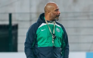 Le sélectionneur de l'équipe du Nigeria, veste verte et bleu marine, observe l'entraînement avec concentration, sur fond de stade flou.