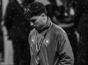Moroccan player wearing a national team jacket and medals, looking down in disappointment under falling rain, reflecting a poignant moment.