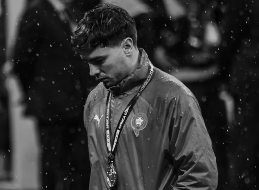 Moroccan player wearing a national team jacket and medals, looking down in disappointment under falling rain, reflecting a poignant moment.