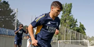 Un joueur du Real Madrid court pendant l'entraînement, vêtu de la tenue marine du club. Des arbres verts et des cages d'entraînement sont visibles en arrière-plan.