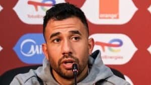 Moroccan player at a press conference, wearing a grey hoodie, speaking with a microphone in front of branded backdrop, focused expression.