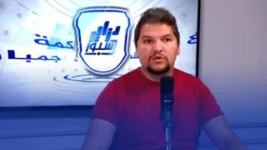 Un analyste sportif s'exprimant dans un studio, portant une chemise rouge, avec un micro et un fond arborant un logo et un texte en arabe.