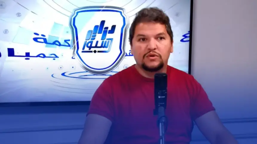 Un analyste sportif s'exprimant dans un studio, portant une chemise rouge, avec un micro et un fond arborant un logo et un texte en arabe.