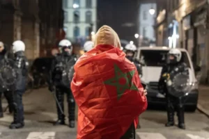 A supporter wrapped in the Moroccan flag stands facing a line of police in riot gear on a dimly lit street, reflecting tension after a match.