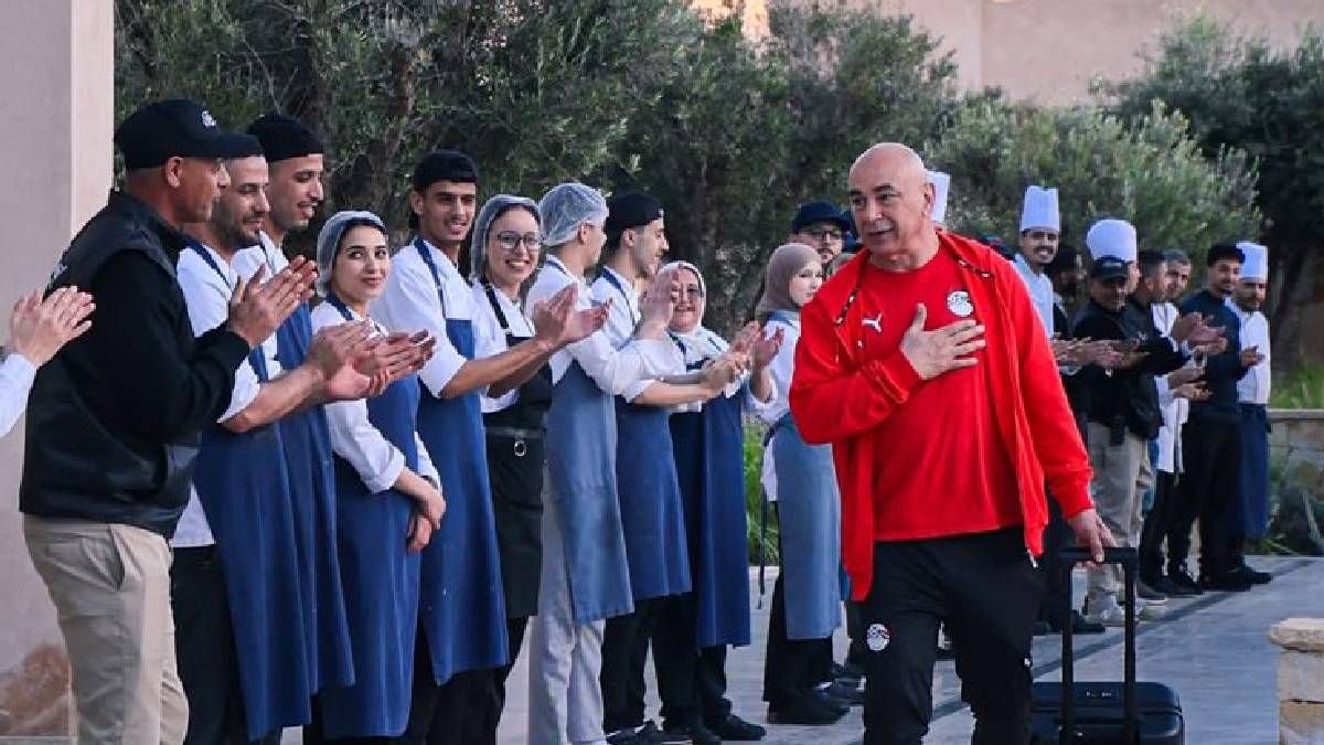 Egyptische coach in een rode trainingsoutfit loopt voorbij een juichende groep keukenpersoneel in schorten, met de handen samen in waardering.