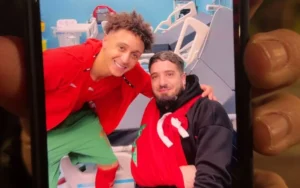 Eliesse Ben Seghir pose avec un supporter vêtu de l'équipement de l'équipe nationale marocaine, tous deux souriant dans un cadre hospitalier, célébrant la camaraderie footballistique.