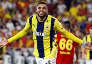 En-Nesyri celebrates joyfully wearing the yellow and navy Fenerbahçe kit, surrounded by enthusiastic fans in a vibrant stadium.