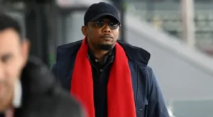 Samuel Eto'o walks confidently, wearing a black jacket and a bright red scarf, with sunglasses, in a bustling urban setting.