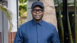 Een man in een marineblauw shirt en zwarte pet staat in een tropische omgeving, glimlacht zelfverzekerd tegen een achtergrond van groen.