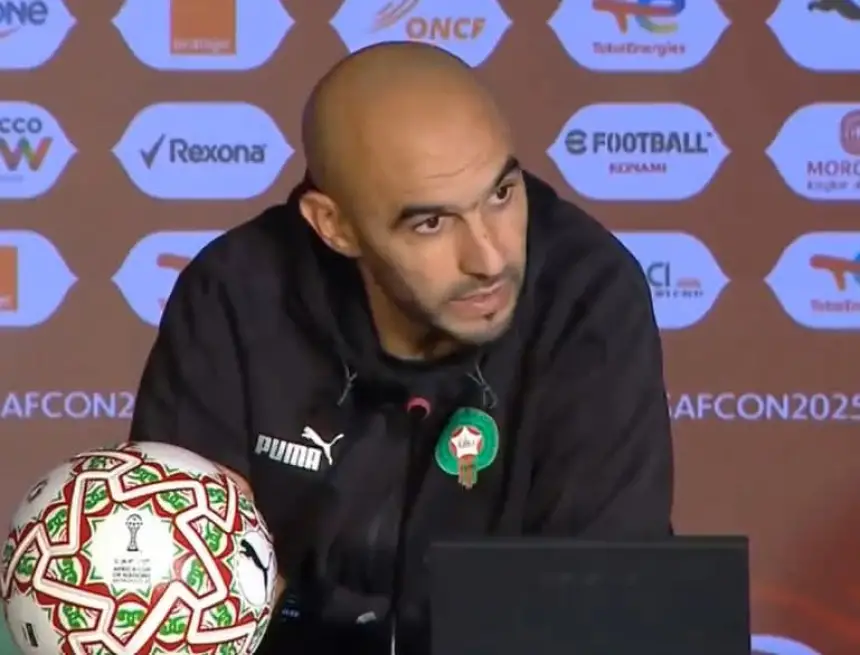 L'entraîneur de football marocain s'exprime lors d'une conférence de presse, avec un ballon décoré de la CAN et le badge de l'équipe visibles sur la table.