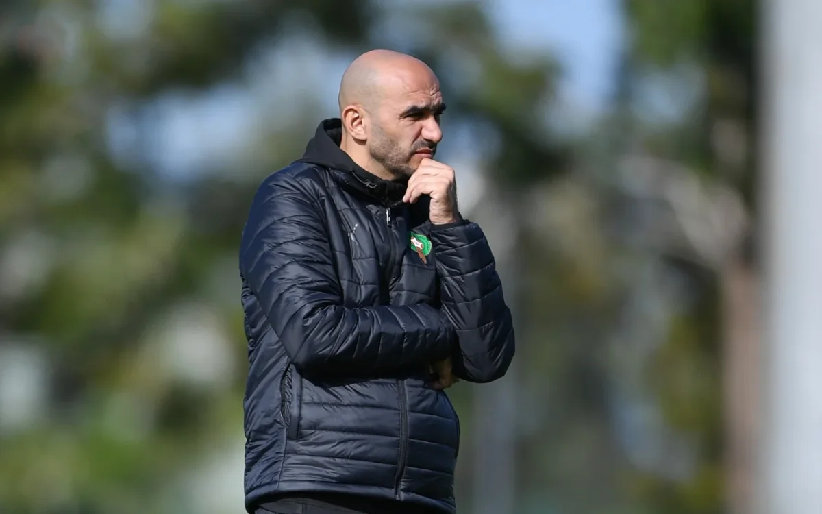 Walid Regragui, vêtu d'une doudoune marine arborant l'emblème marocain, réfléchit lors d'une séance d'entraînement, entouré de verdure.